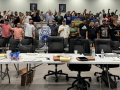 A group of 50 people stand behind a bargaining table with fists raised. 