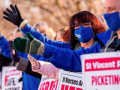 Woman in blue coat facing sideways with hand up among many picketers with hands up, who are holding signs in support of St. Vincent striking nurses