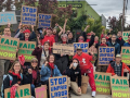 A group of workers and supporters hold cardboard signs reading Stop Unfair Labor Practices and Fair Contract Now. Many are wearing red bandanas.