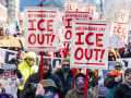 View of a section of a giant crowd marching, on a bright sunny day, bundled against extreme cold. Prominent picket signs that look screen-printed or stenciled say "Neighbors say ICE out!" in crisp red on white background.