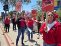 Teachers in red picket alongside a road with “Fully Staff Our Schools signs”