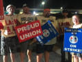 Four men and one woman wearing Machinists tabards and holding strike signs face the camera at night with plant lights behind them.