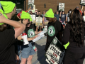 Several people in high-viz green stocking caps gather around a sheet of paper, pickets in the background have signs saying ‘no contract, no coffee’