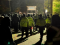 A Sysco truck faces off against a crowd of people, backs to the camera. A few wear yellow jackets that say "Police" or "Sheriff" on the backs; most do not. Around them it is still very dark out, but the central paved area is lit from above.