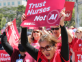 Women workers in red scrubs rally and chant, outdoors on a sunny day. They hold printed NNU signs. The woman most visible, in big sunglasses, holds a printed sign reading "Trust nurses, not A.I."