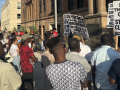 A group of workers, mostly Black, face an official-looking building with signs.