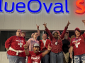 A group of 7 workers look excited and raise fists in front of a “BlueOval SK” sign