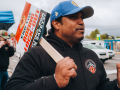 Picketer in IAFF hoodie and ballcap stands on a sidewalk, speaking and gesturing. Over his shoulder is a printed picket sign that says "End the Boeing Lockout. Boeing Firefighters - Fighting for Your Safety!"