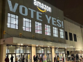 Warehouse lit up against dark sky. Directly under the "Amazon" logo the message "Vote yes!" is projected on the wall. A few people are visible coming and going near the doors below.