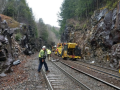 Workers in reflective yellow jackets work outdoors on railroad tracks, with heavy machinery, surrounded by rocky cliffs and forest