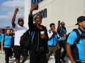 Workers, some in blue vests, march outside along an Amazon building. Some are holding their phones aloft, recording. Many are Black. They look like they are striding confidently and chanting. 