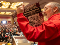 Seen from the side/back, Shawn Fain in a red hoodie holds up a battered purple copy of A Troublemaker's Handbook. Arrayed in front of him can be seen a huge seated crowd in the ballroom for the closing plenary.