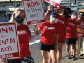 Six picketers in red NUHW T-shirts stand on outdoors along a sidewalk. Most are masked. Some carry handmade signs that say "Honk for mental health" and "honk for patient care." Others carry printed signs that say "Fair contract now" and "Patient health not corporate wealth."