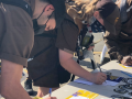 Three people in UPS uniforms and masks bend over a table of campaign literature, signing cards