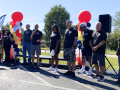 About a dozen adults and a couple children, white and Black, men and women, stand mostly silhouetted in bright sun. Most wear black union T-shirts.They're outdoors, on pavement in front of grass. One holds a mic. Some hold printed signs that say, "IUE-CWA, Better Wages." There are also two columns of balloons, each a spiral design in white, red, yellow, and black.