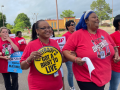 Women, mostly Black women, march outdoors in red T-shirts that say "Organize" with a telephone logo. Most prominent in the foreground are three women who have linked elbows and are smiling big. The woman on the left has a yellow round sign that says "Everybody's got a right to live." The woman in the middle has blue hair. The woman on the right is holding a light blue sign we can't see, but it probably matches signs visible behind which say "CWA on strike against Maximus." One woman is smoking.