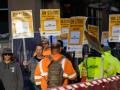 Picketers in high-visibility garb file alongside a plastic barrier. Printed signs read "ON STRIKE FOR FAIR WAGES AGAINST GLY CONSTRUCTION"