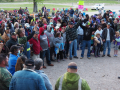 A crowd of people forms a big circle outdoors. One person stands in the middle speaking.