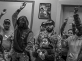 Black and white photo shows workers raising their hands, some holding kids, indoors with a small Rosie the Riveter poster on a nearby wall.