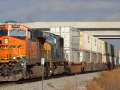 A freight train passes along a track under an overpass. The orange engine is labeled BNSF. It is pulling many white freight cars.