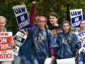 Six workers march carrying printed "UAW on strike" signs and wearing rain ponchos. One smiles, and others look determined; one person wearing two long braids punches their fist high in the air. They are outdoors on a green campus.