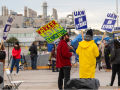 Workers in heavy coats stand around picketing in front of a big gray factory. One neon-colored handmade sign says "Worker-led EV transition" with a lightning bolt. Other signs are printed and say "UAW on strike" or "UAW, Stand up, Record Profits, Record Contracts" 