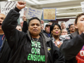 Close-up of a diverse crowd standing, clapping,shouting, raising fists. T-shirts in front row say "save our pensions" and "we are one." Banner on wall behind them says "Machinists District [751], Keep America Working." Handmade signs say things like "No no no, don't sell your soul," and "Thanks IAM, you're standing up for all of us."