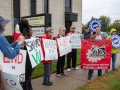 People with signs stand on grass outside an office building. The man speaking holds a red banner that says, "Solo organizados conquistaremos nuestros derechos laborales. Casa Bajia Obrera." Others listening hold printed blue UAW logo signs and hand-written signs that say "Shame on VU" or "End the blacklist." 