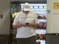 A person in a T-shirt with the message "#RidinwithRico" and a Mercedes logo hat looks down at their phone. They are standing in front of a vending machine.