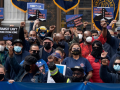 A diverse crowd, all masked, some with fists in the air, outdoors on steps. Printed signs read: "Transit can't work from home," "Hazard pay for transit workers," and "Call us heroes, pay us zeroes" 