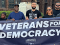 A group of veterans stand behind a blue banner that reads Veterans for Democracy.