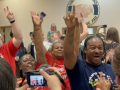 Several people raise their fists indoors, they look excited and happy.