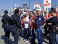 a group of picketers face three police officers. They are holding strike signs. A refinery truck is to the left.