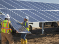 Three solar workers in yellow vests lift a large solar panel into place on a solar array.