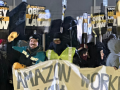 A crowd of workers stands outdoors in dim light. A smiling person with raised fist is one of several holding a handmade banner--the part visible says "Amazon Workers." Many people in crowd hold printed Teamster-logo picket signs that say: "Amazon: Obey the Law." Everyone is wearing coats with big hoods or warm hats; it looks cold out. Someone in lower left has a Staten Island T-shirt and keffiyeh