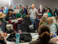 In a hotel conference room, one person stands and talks, while many seated at circular tables or standing against a wall listen thoughfully.