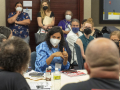 People are gathered around a table wearing masks, a woman with dark hair speaks and gestures in the middle.