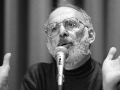 A bearded man speaks at a microphone with his hands wide open