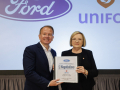 A man and a woman in suits stand holding a binder together in front of a screen that has Ford and Unifor logos projected on it.