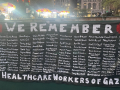 A black banner with white letters lists 160 health care workers killed in Gaza. It is titled “We Remember”.