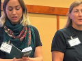 Two women in dark t-shirts stand side by side, one reading from a thick booklet