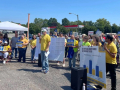 A group in a parking lot in yellow t-shirts listen while one man speaks through a microphone