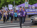 Workers in purple SEIU Local 49 T-shirts march across a street. Some carry picket signs that say SEIU and some other words too small to read. A large purple banner says "We stand for our patients." The person in front is talking into a bullhorn and others appear to be chanting. 