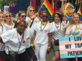 Nurses in white scrubs with rainbow flags and accessories; one carries a sign "I Love My Trans Patients" with the trans flag (pastel blue, pink, and white stripes)