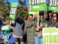 Women stand on a sunny day, wearing big jackets, holding green printed ONA picket signs that say "Unfair Labor Practice Strike" and "Nurses on Strike." A handmade sign visible in the background says "Burnout caused this turnout."