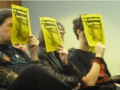 Protesters sitting in a row in a meeting hold up yellow posters with a picture of slain worker Juan Baten.