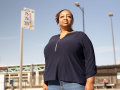 Black woman looks into the distance with a serious, determined expression. Behind her is a bus stop sign and a blue sky.