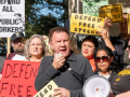 Tom Alter, a white man with a short haircut, squints into the sun and speaks into a megaphone, He is surrounded by a tightly packed crowd, diverse in race and gender, holding signs with slogans like "Defend Free Speech," "Defend All Public Workers," and "Reinstate Tom Alter."