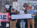 Eight people outside face the camera with signs, including “Hire Genesis” and a UE banner. 