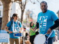 Two men in blue shirts that say “UPTE” play snare drums while others march in the background.